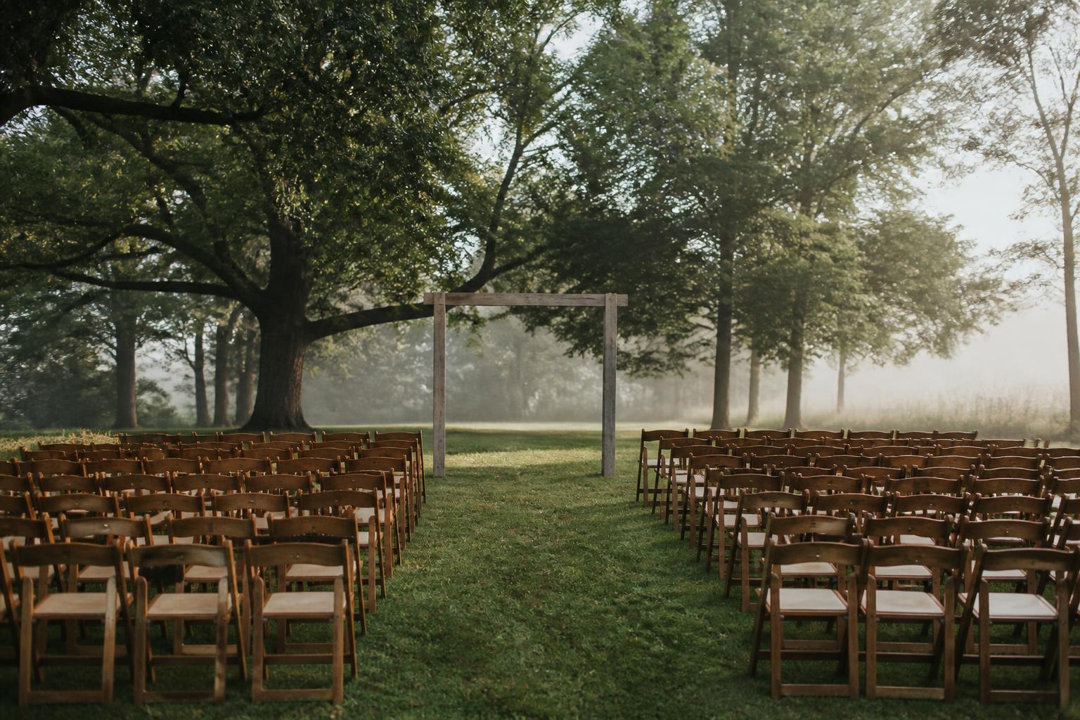 Rows of simple wooden chairs facing a plain wooden arch on a lawn beneath mature trees in early morning light
