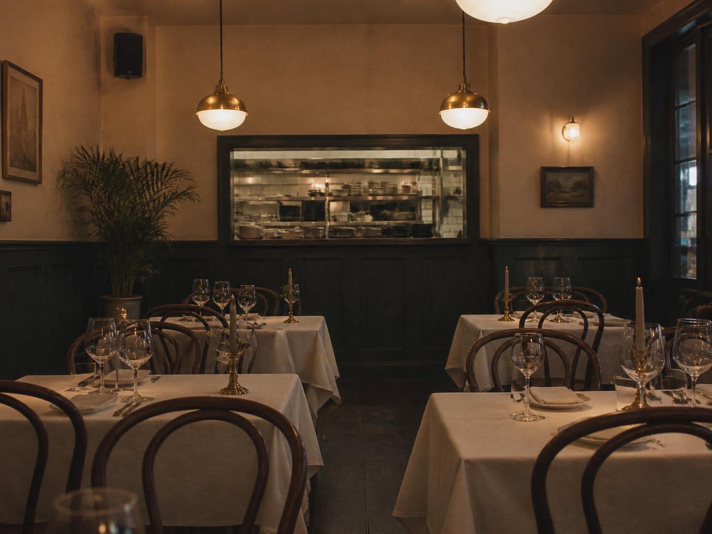 A small restaurant dining room set before service, with brass pendant lights and an open kitchen pass